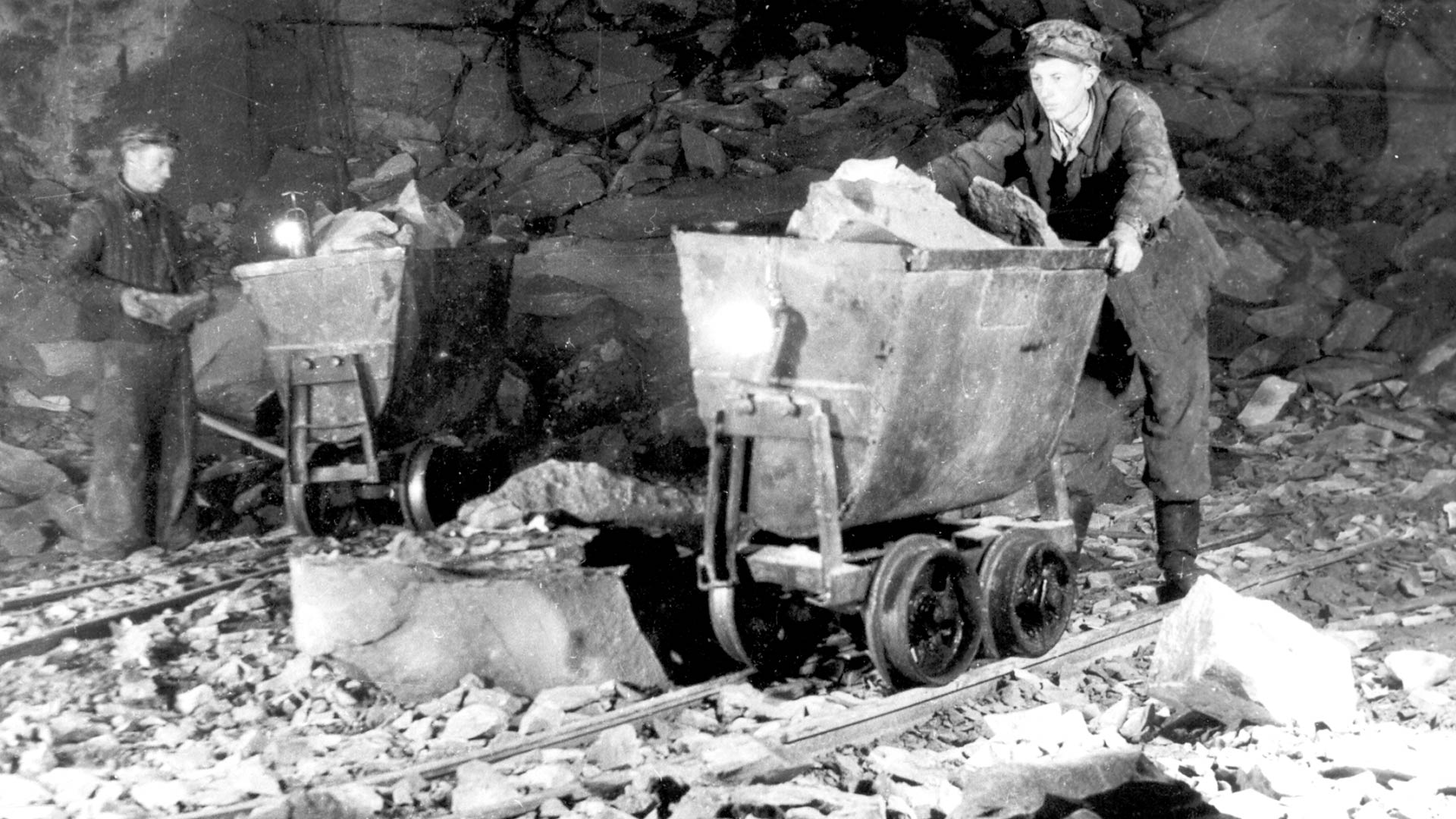 Historic black and white image of an old smelting complex. A man is seen wheeling a cart. 