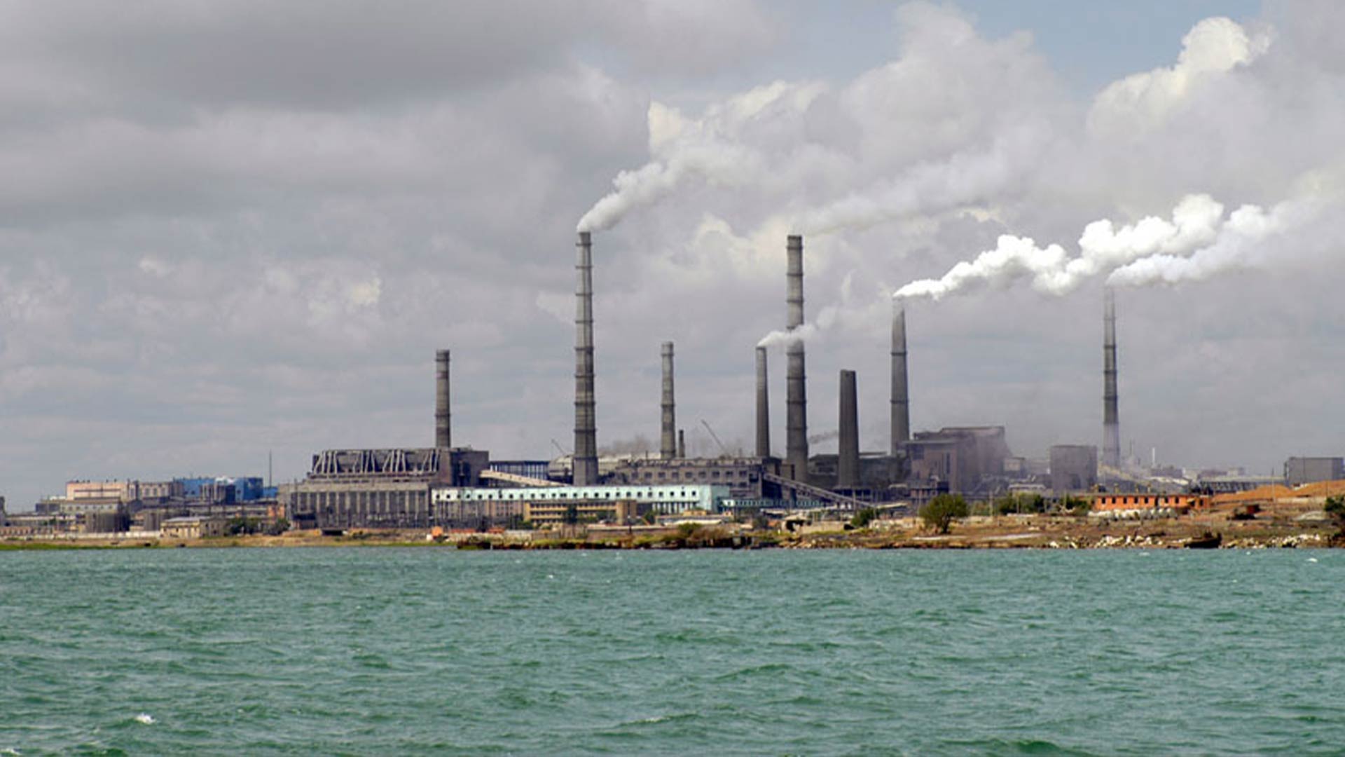 A distant image taken from across a body of water. On the other side, buildings and factories are seen with smoke coming from multiple long chimneys. 