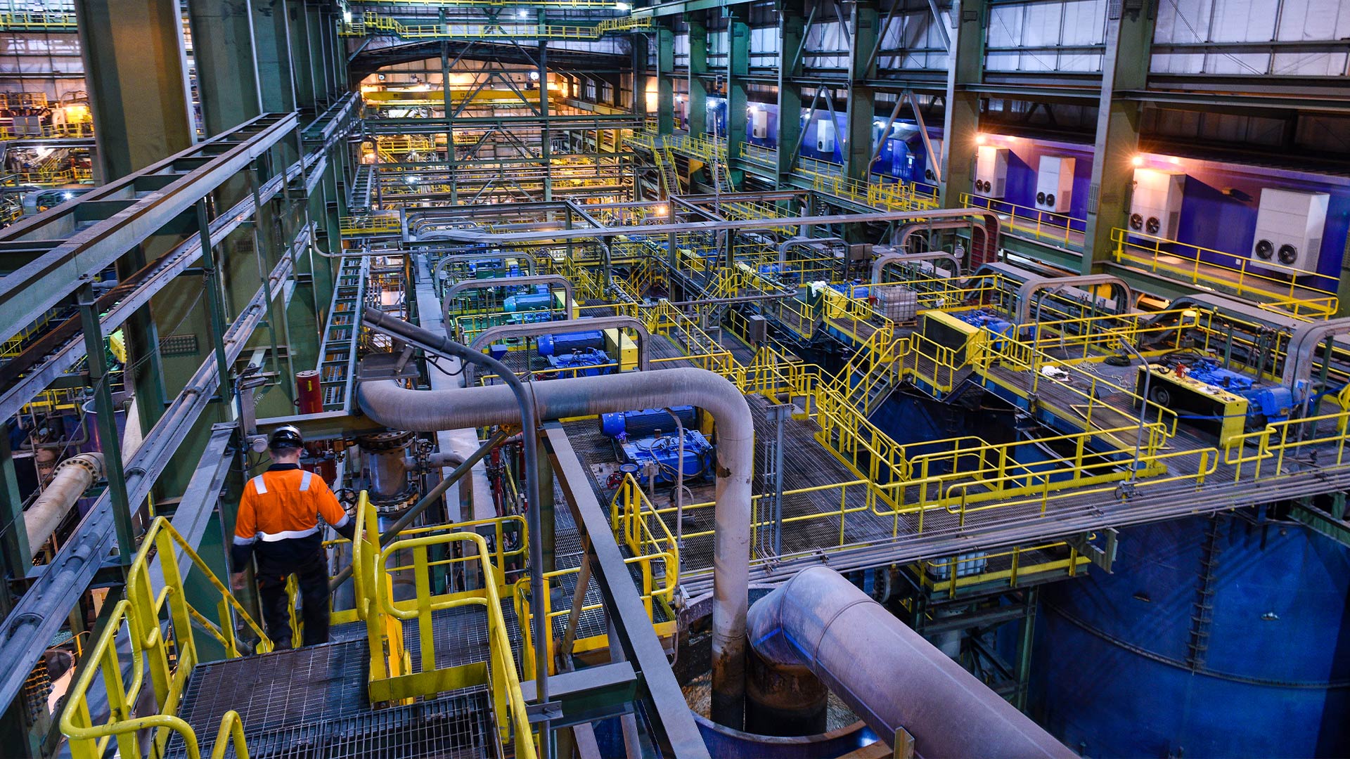 Тау-кен өндірісінің ішінде. Сол жақта баспалдақтан түсіп келе жатқан жұмысшы көрінеді. Кең кеңістікті қамтитын көптеген күрделі түтіктер мен қоршаулар бар.