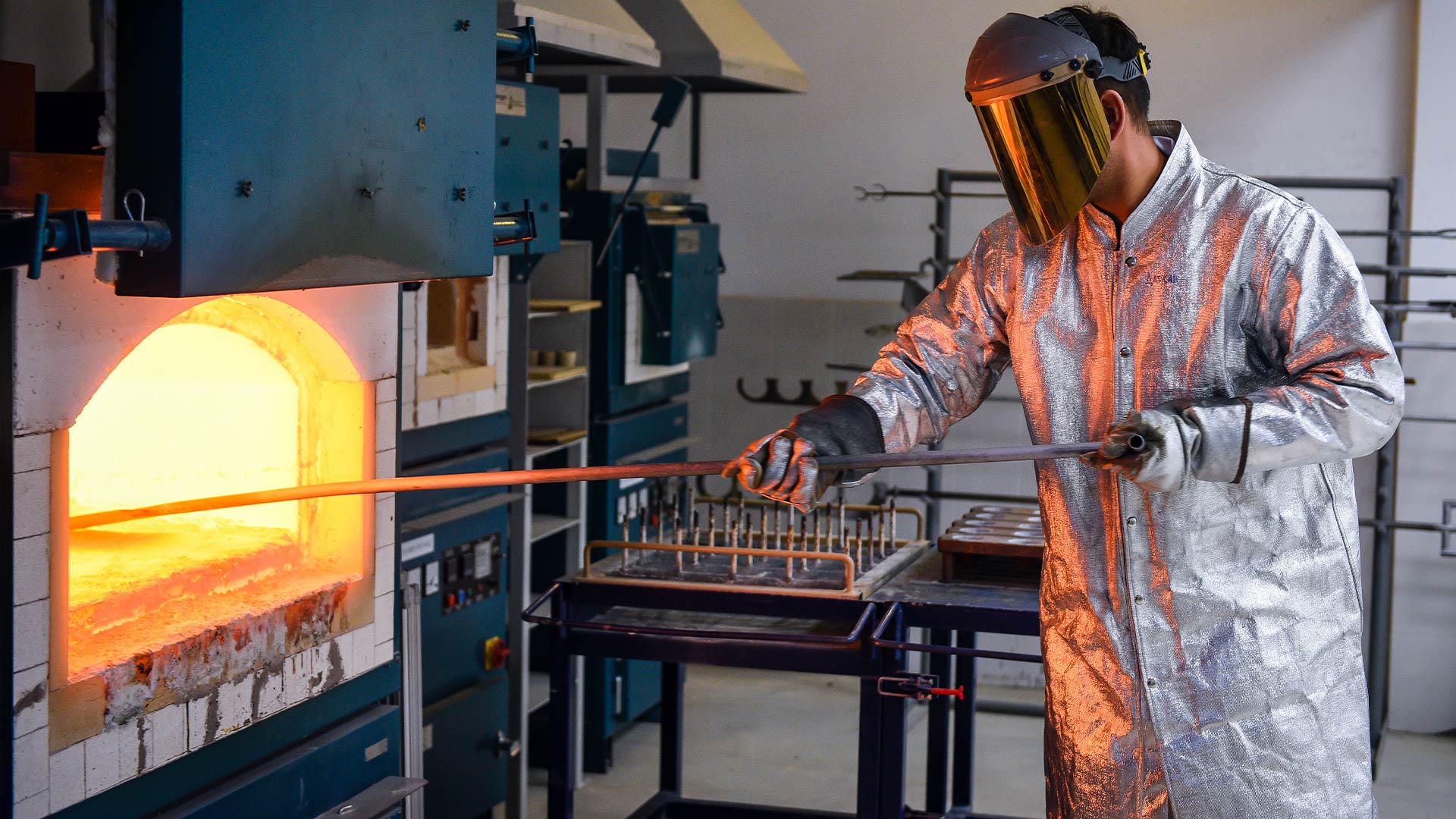 A worker is seen in full safety headwear and clothing putting a rod into a furnace. 