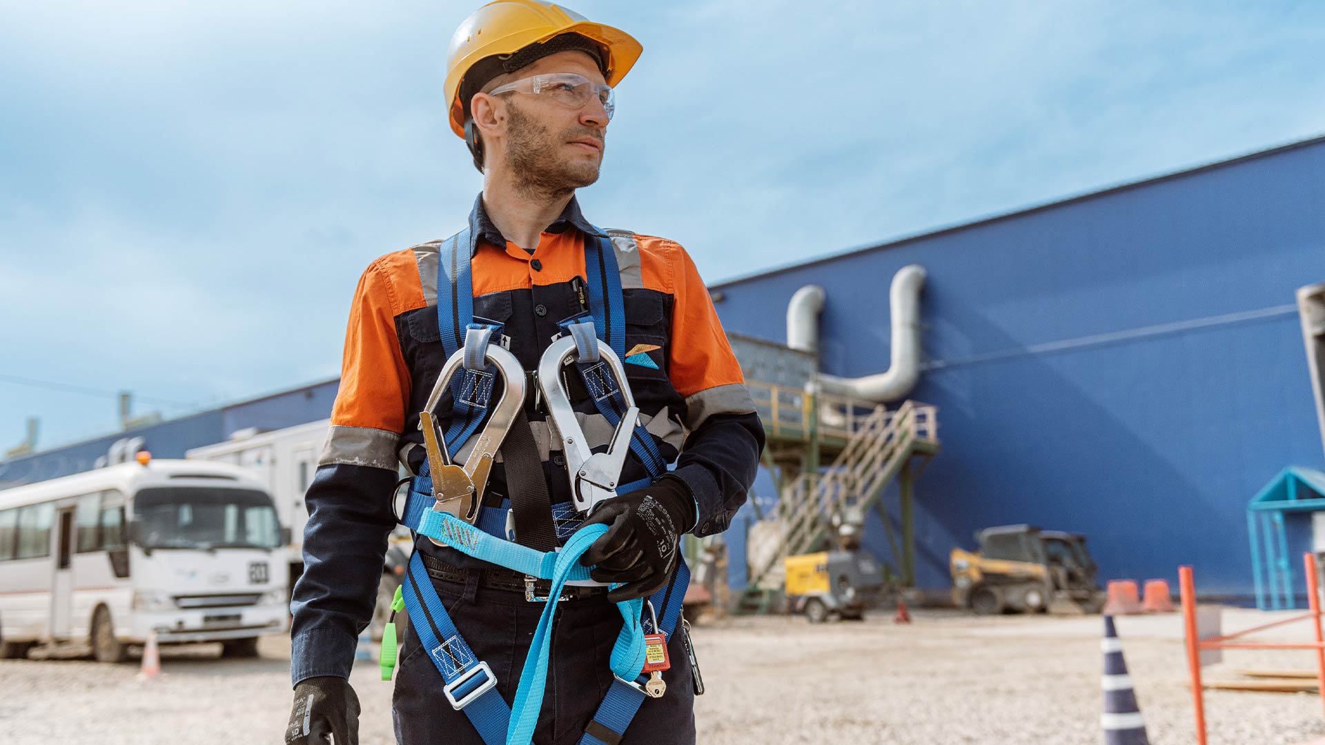 Worker standing in safety gear looking into the distance. Behind him is a blue building and light blue skies. 