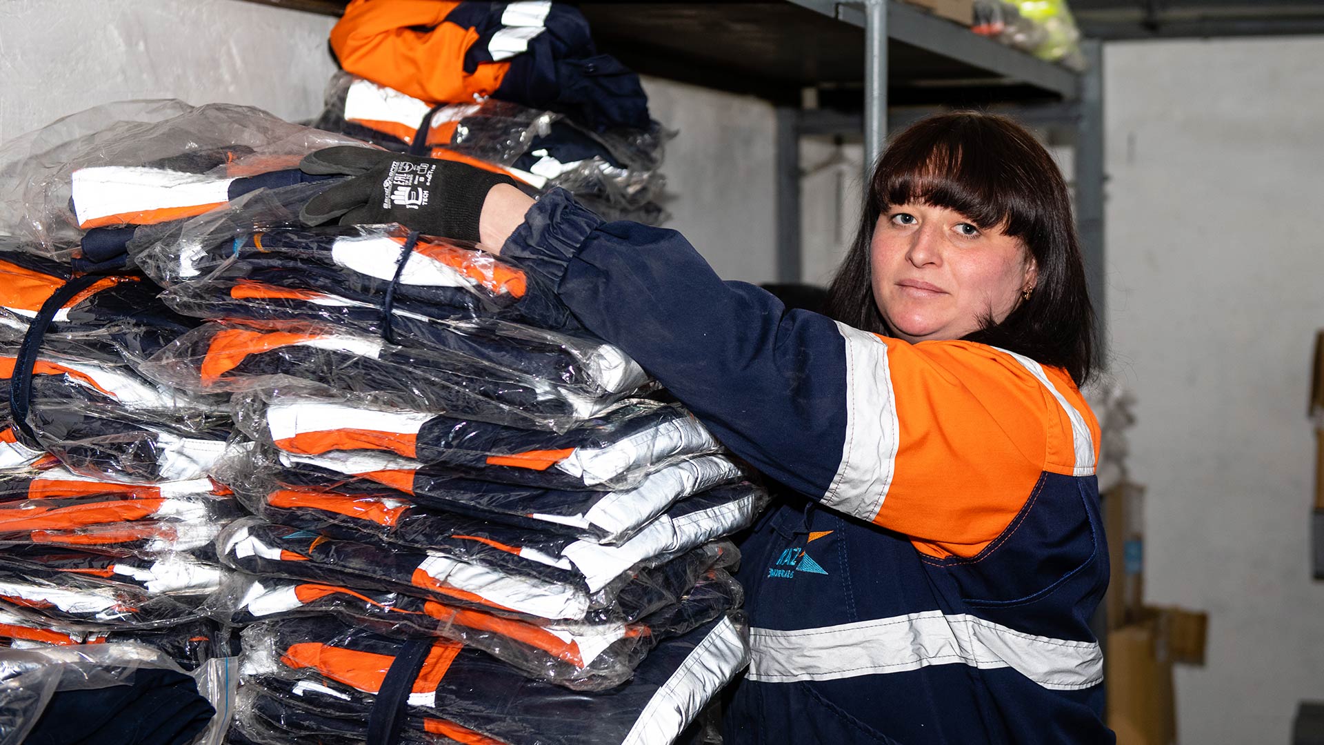 Қауіпсіздік құралын киген адам камераға қарап тұр. Артқы жағында техника көрінеді.