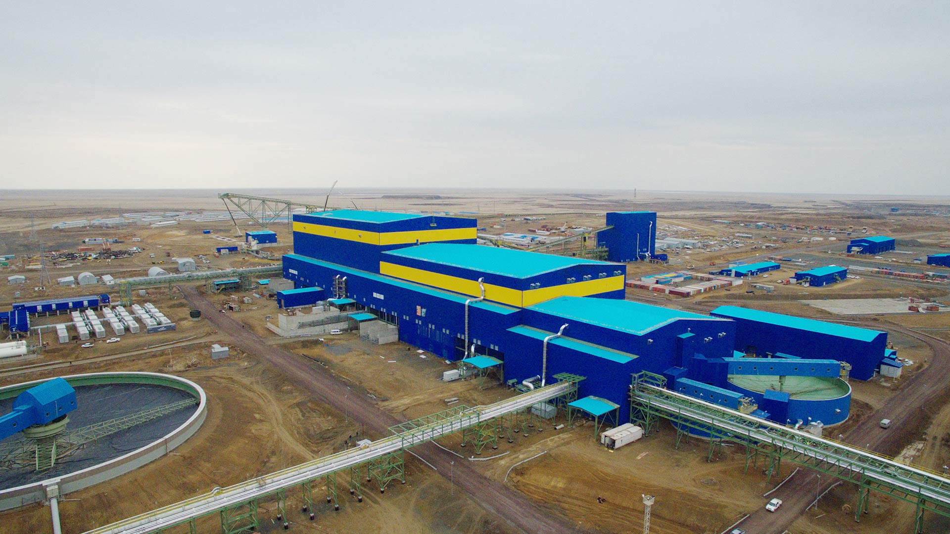 A vast industrial complex with large blue and yellow buildings under a cloudy sky. Surrounding barren landscape with machinery and pipes conveys an industrial tone.