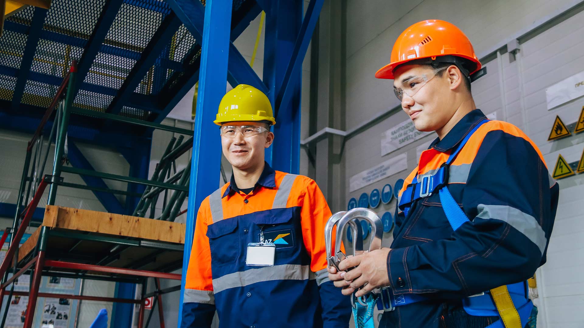 Two workers in bright safety gear, including helmets and goggles, stand in a warehouse. One holds coiled cables, conveying teamwork and readiness.